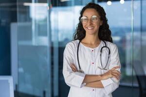Portrait of a beautiful Latin American female doctor, doctor in working in a modern clinic office, standing by the window in a beggar's robe, with crossed arms smiling and looking out the window photo