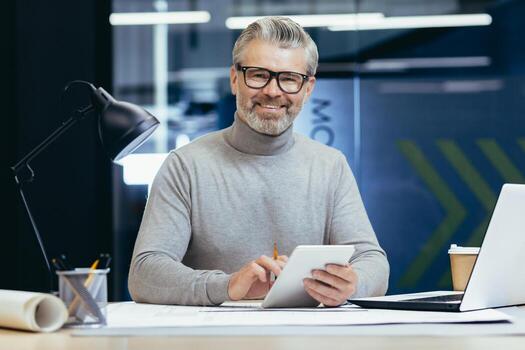 Portrait of senior architect, gray haired designer smiling and looking at camera, man mock up plan use laptop and tablet computer at work inside studio office. photo