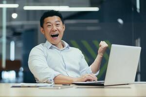 Successful and happy young Asian man, businessman, rejoices, shows yes with his hand, looks at the camera. Made a contract. Won money. Sitting with a notebook at the table in a modern office. photo