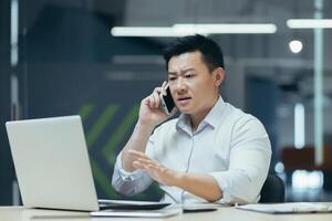 Worried and nervous young Asian man talking on the phone in the office, looking at the laptop, solving problems, questions. photo