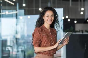 Young and successful female programmer, portrait of female engineer with tablet computer startup worker working inside office building using tablet for testing applications smiling looking at camera. photo