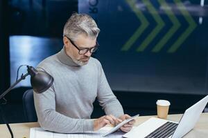 Serious thinking gray-haired businessman working inside office with laptop, senior man using tablet computer browsing internet pages reading online news. photo