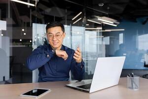 Cheerful and smiling online customer support tech worker, Asian man with call headset smiling and looking at webcam, working on helpline and consulting customers, using headset. photo