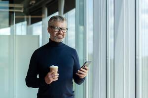 A senior male businessman stands in an office center near a window and uses the phone. He holds a cup of coffee in his hand. Went on a break, rest at work, coffee break. He looks at the camera smiling. photo