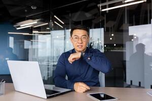 Successful businessman inside office, using app on smart watch to communicate, asian man giving voice commands to artificial intelligence through watch, boss working sitting with laptop. photo