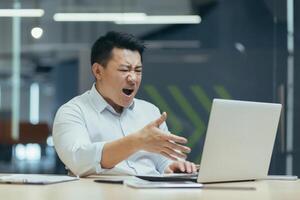 Angry and dissatisfied with the result of the work, a young Asian male businessman sitting in the office in front of a laptop and shouting at the screen on a call. photo