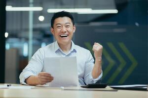 Portrait of successful asian businessman boss, man smiling and looking at camera, holding letter and document in hands, happy with success holding hand up gesture of triumph and victory photo