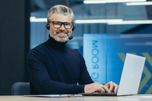 Portrait of mature gray haired businessman, man in glasses smiling and looking at camera, senior boss with headset for call using laptop, executive working inside modern office building. photo