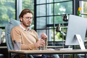 Sick and overtired office worker at work with a computer, businessman inside the office takes a medical pill and drinks water, a blond man in casual clothes, a sweater with a shirt. photo
