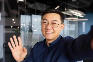 Asian man talking on call using online communication app on phone, businessman in office taking selfie and smiling at camera, man greeting waving hand while talking with friends. photo