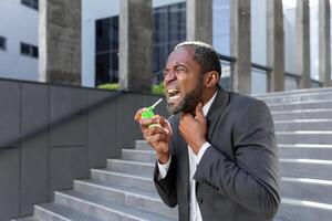 An older African American man in a suit clears his throat from pain and inflammation while standing outside an office center. photo