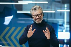 Senior gray haired businessman talking on call, using headset investor in glasses smiling and looking at camera, webcam view man working inside modern office building. photo