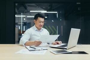 Office work. A young Asian man is working on a laptop and documents while sitting at a table. photo