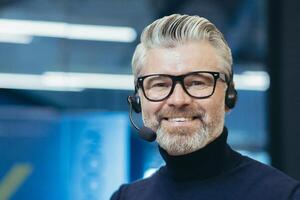 Portrait of a gray-haired handsome man in glasses and a headset with a microphone. Speaker, business trainer, online conference. Looking at the camera, smiling. photo