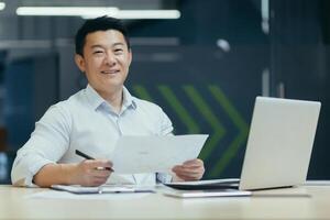 Office work. Portrait of a young Asian man working on a laptop and documents while sitting at a table. He looks at the camera with a smile. photo