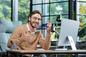 A young man poses for the camera, advertises a medicine for asthma and allergies. He sits in the office at a table with a computer, holds an inhaler in his hand, shows super with his hand, smiles. photo