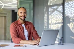 Smiling man working on laptop at desk in modern office, showcasing productivity. Table holds smartphone and pen holder, reflecting efficiency and work environment. Bright, professional setting. photo