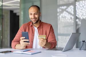 Smiling man holding smartphone and credit card while online shopping at office desk. Engaged in digital purchasing with phone, displaying convenience and modern technology. photo