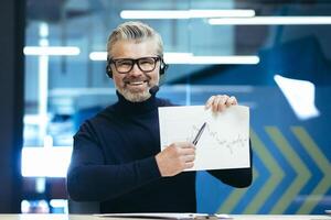 A man in headphones with a microphone. Sitting in the office, holding graphics, a project in his hands and pointing to the camera with a pen. Conducts business meeting, web conference, smiles. photo