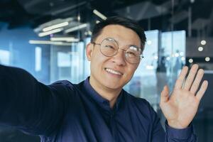 Smiling and cheerful asian businessman boss investor in glasses using smart phone for call, looking into phone camera and waving hand gesture of greeting, talking with colleagues inside office. photo