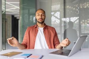 Man practicing meditation in office setting, sitting at desk with closed eyes. Laptop and notebooks indicate work environment. Promotes mindfulness, stress relief, and focus during work. photo