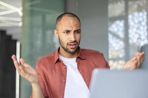A perplexed office worker sits at his desk, gesturing in confusion while looking at his computer screen. The scene suggests a problematic virtual meeting or unexpected document issue. photo