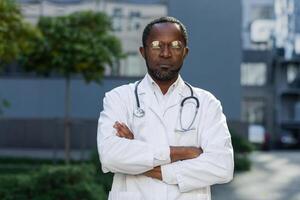 Portrait of serious thinking african american doctor, man in glasses and medical coat with stethoscope looking at camera with crossed arms outside modern clinic. photo