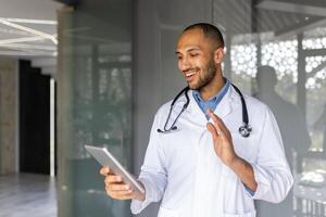 Professional doctor wearing stethoscope having friendly call on tablet. Smiling and gesturing, showcasing modern healthcare communication and digital interaction in medical environment. photo