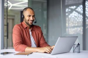 Smiling professional wearing headset works on laptop in modern office setting. Engaged in online meeting using phone technology. Notebook and pen on table indicate readiness and focus. photo
