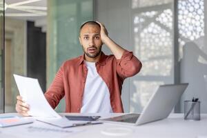A professional looks worried while examining documents in a modern office setup, showing stress or confusion. The setting includes a laptop and charts, highlighting a business or financial context. photo
