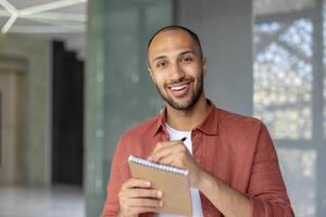 A confident man with a friendly smile is holding a notepad and pen inside a modern space, showcasing a positive and productive atmosphere. photo