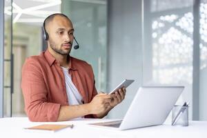 A young professional man is using a smartphone and laptop at his desk, wearing a headset. The scene suggests a remote work environment, highlighting technology use and communication. photo