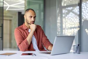 Thoughtful man with laptop in modern office. Professional analyzing work strategy. Wearing casual attire, promoting focused approach to tasks. Phone and notebook nearby for efficiency. photo