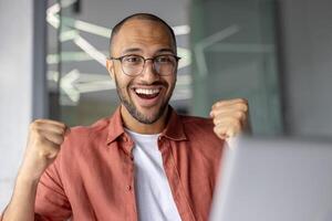Energetic young man with glasses celebrating success with fists raised and joyful expression in modern office environment, conveying motivation and achievement. photo