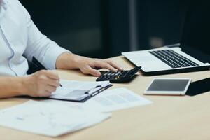 Close-up photo. Male hands in a white shirt at the table in the office with a tablet, laptop, calculator. They write with a pen on paper, sign documents, work with accounts and finances, reports photo