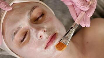 professional cosmetologist applies a white mask on the face of a female client with a brush, close-up photo