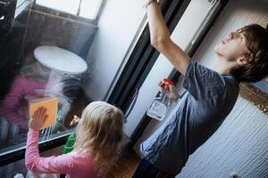 Child cleaning a window while an adult supervises in an indoor setting during a sunny day photo