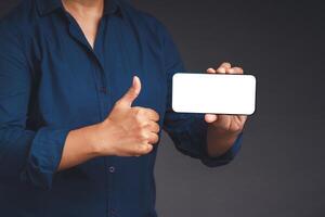 Hand holding a horizontal mobile phone with a blank white screen and showing thumbs up while standing on a gray background photo