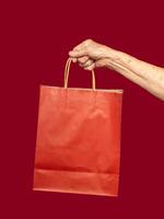 A Close-up of a senior woman's hand holding a red paper bag with a handle against a red background photo