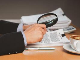 Close-up of businessman hand holding a magnifying glass looking through documents while sitting in the office photo