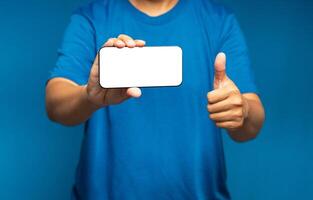 Hand holding a horizontal mobile phone with a blank white screen and showing thumbs up while standing on a blue background photo