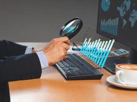 Businessman looking at statistical information comparing data in charts graph with a magnifying glass on a virtual screen while sitting in the office photo