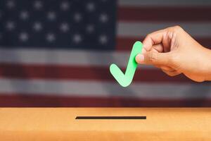 Hand holding a green check mark symbol overhead the voting box at place election with an American flag background photo