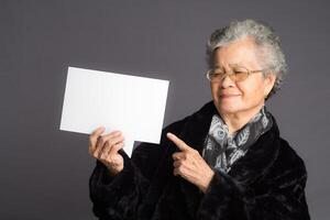 An elderly Asian woman in a sweater with short gray hair holds and points to a blank speech bubble with a smile while standing on a gray background photo
