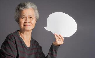 An elderly Asian woman holding a blank speech bubble and looks at the camera with a smile while standing on a gray background photo