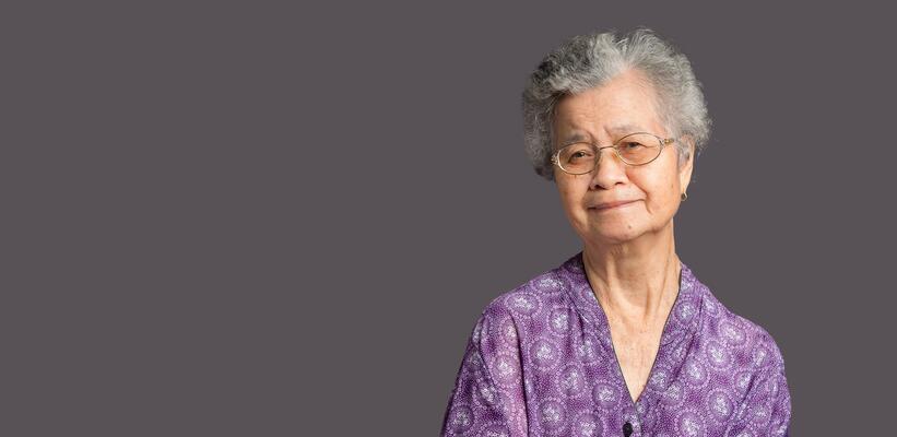 An elderly Asian woman looking at the camera with a smile while standing on gray background