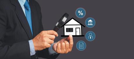A businessman's holding a magnifying glass and looking at a home property while standing over gray background photo