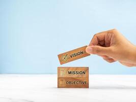 Hand holding a wooden cube with the letters Vision, Mission, and objective on a marble table against a blue background photo