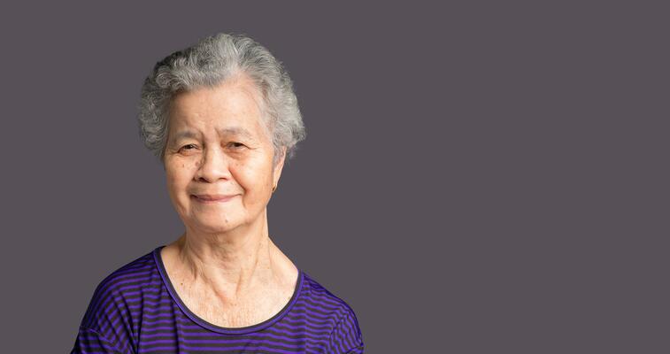 An elderly Asian woman looking at the camera with a smile while standing over a gray background