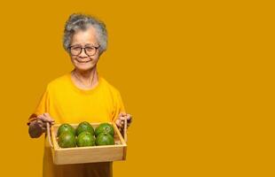 Senior woman with short gray hair holding a wooden tray with avocados and looking at the camera with a smile while standing on a yellow background photo